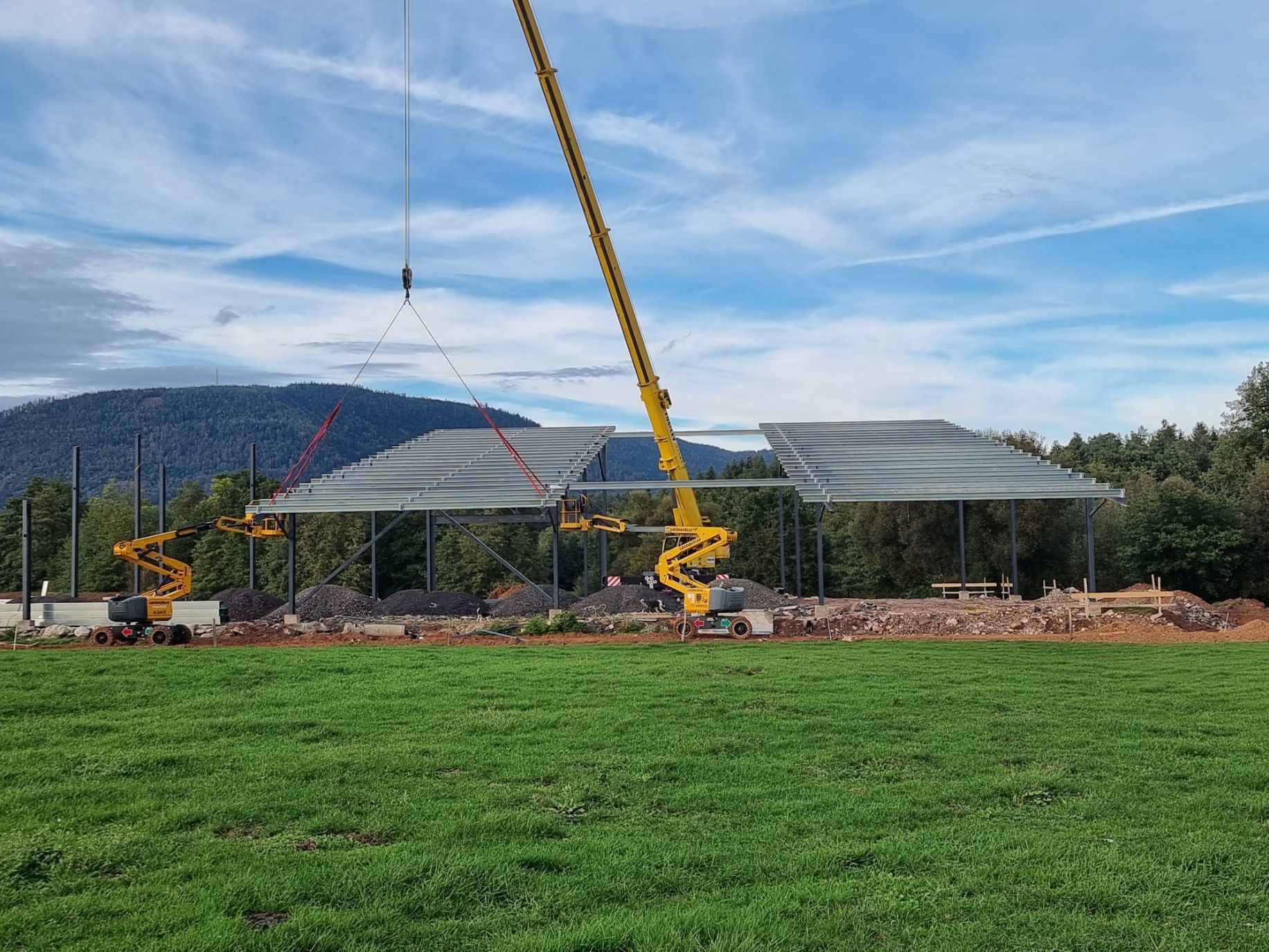Une grue soulève des fermes de toit en acier sur un chantier, avec une pelouse au premier plan et des montagnes en arrière-plan