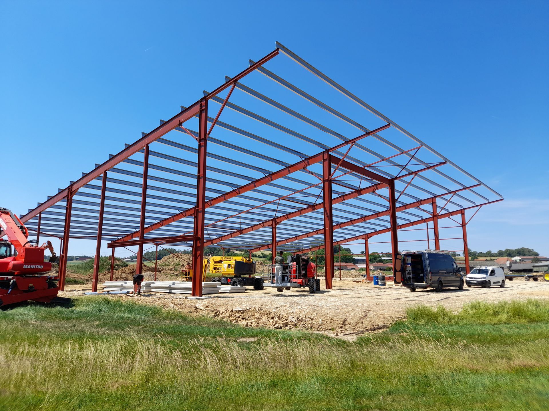 Bâtiment à ossature métallique en construction, avec des ouvriers et des véhicules sur un terrain gravillonné sous un ciel bleu