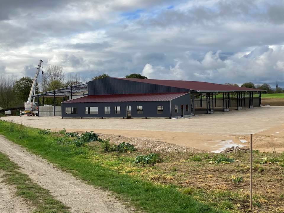 Bâtiment agricole rural aux murs bleu foncé et au toit rouge, près d’un terrain en terre battue dégagé, sous un ciel nuageux