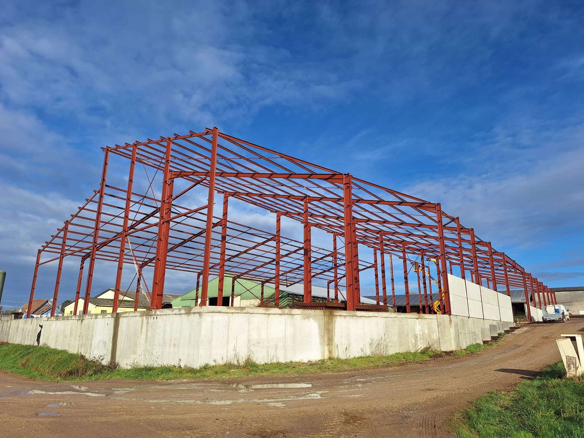 Charpente métallique rouge d’un grand bâtiment en construction sur des fondations en béton, sous un ciel bleu.