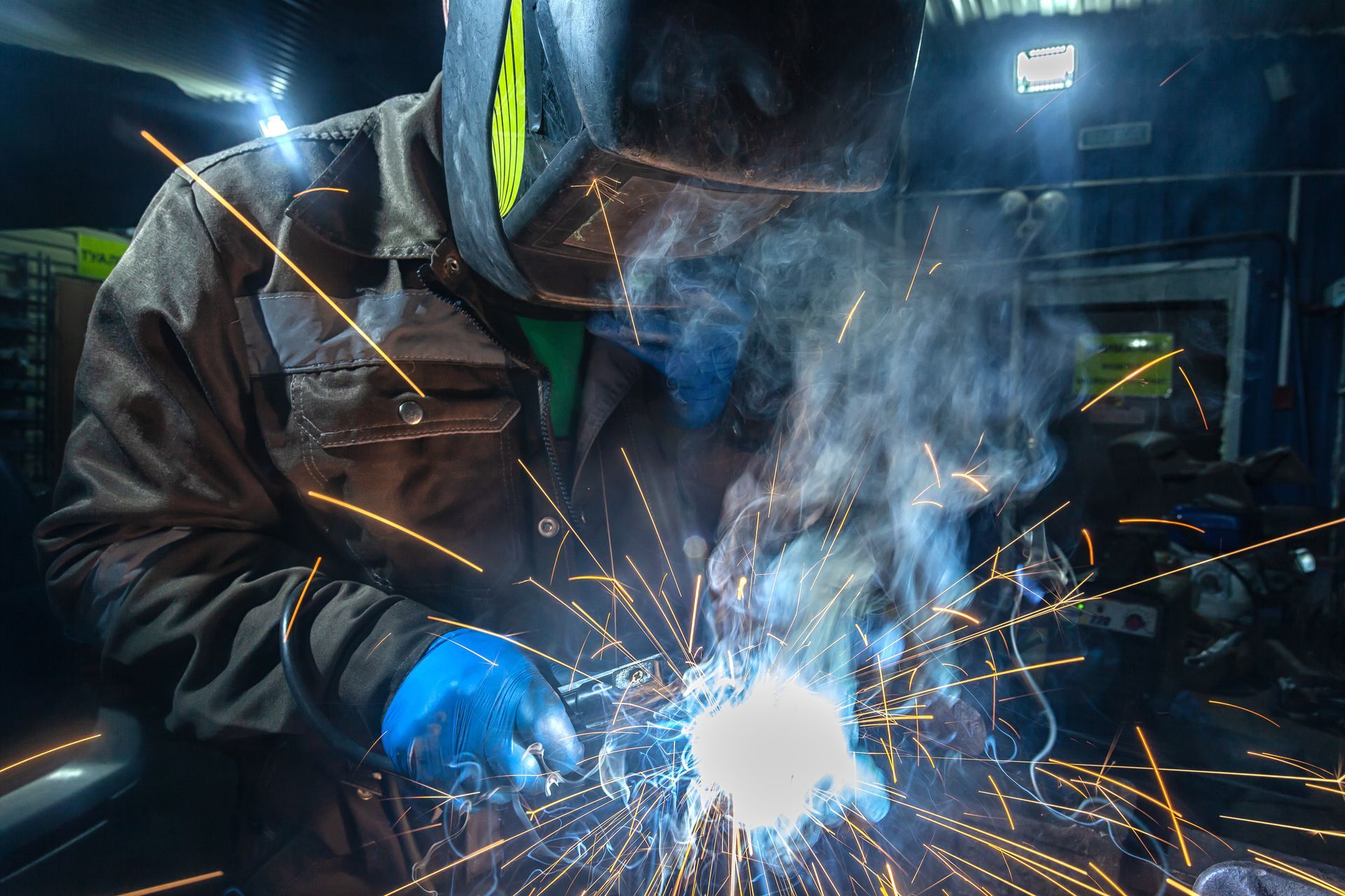 Un soudeur vêtu d’une tenue de protection sombre et de gants bleus fait jaillir des étincelles en travaillant le métal incandescent dans un atelier enfumé