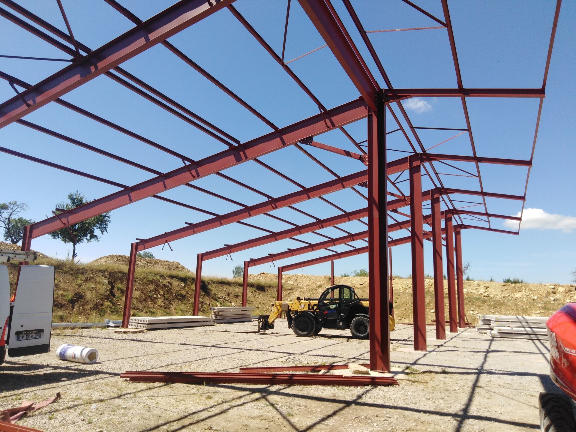 Bâtiment à ossature métallique en construction, avec des poutres rouges, un ciel bleu et un petit véhicule utilitaire à l’intérieur