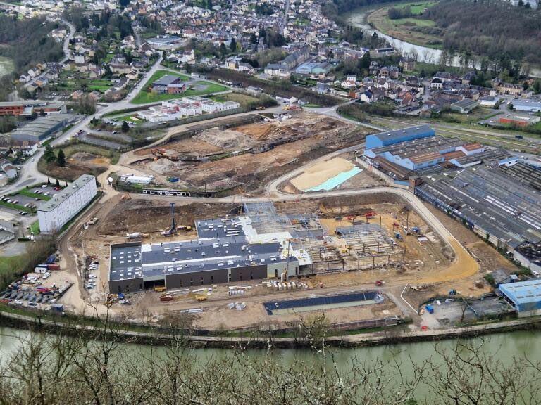 Vue aérienne d’un grand chantier au bord d’une rivière, avec des bâtiments en construction et des travaux de terrassement en cours.