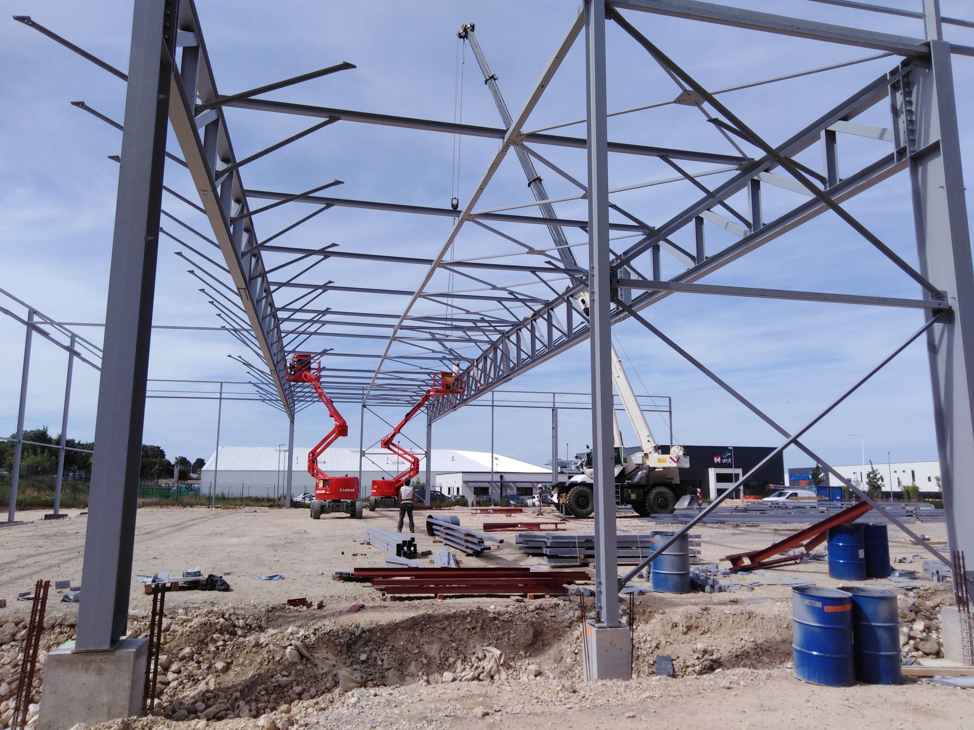 Chantier de construction à ossature métallique, avec des nacelles élévatrices rouges et des ouvriers qui assemblent la charpente du toit