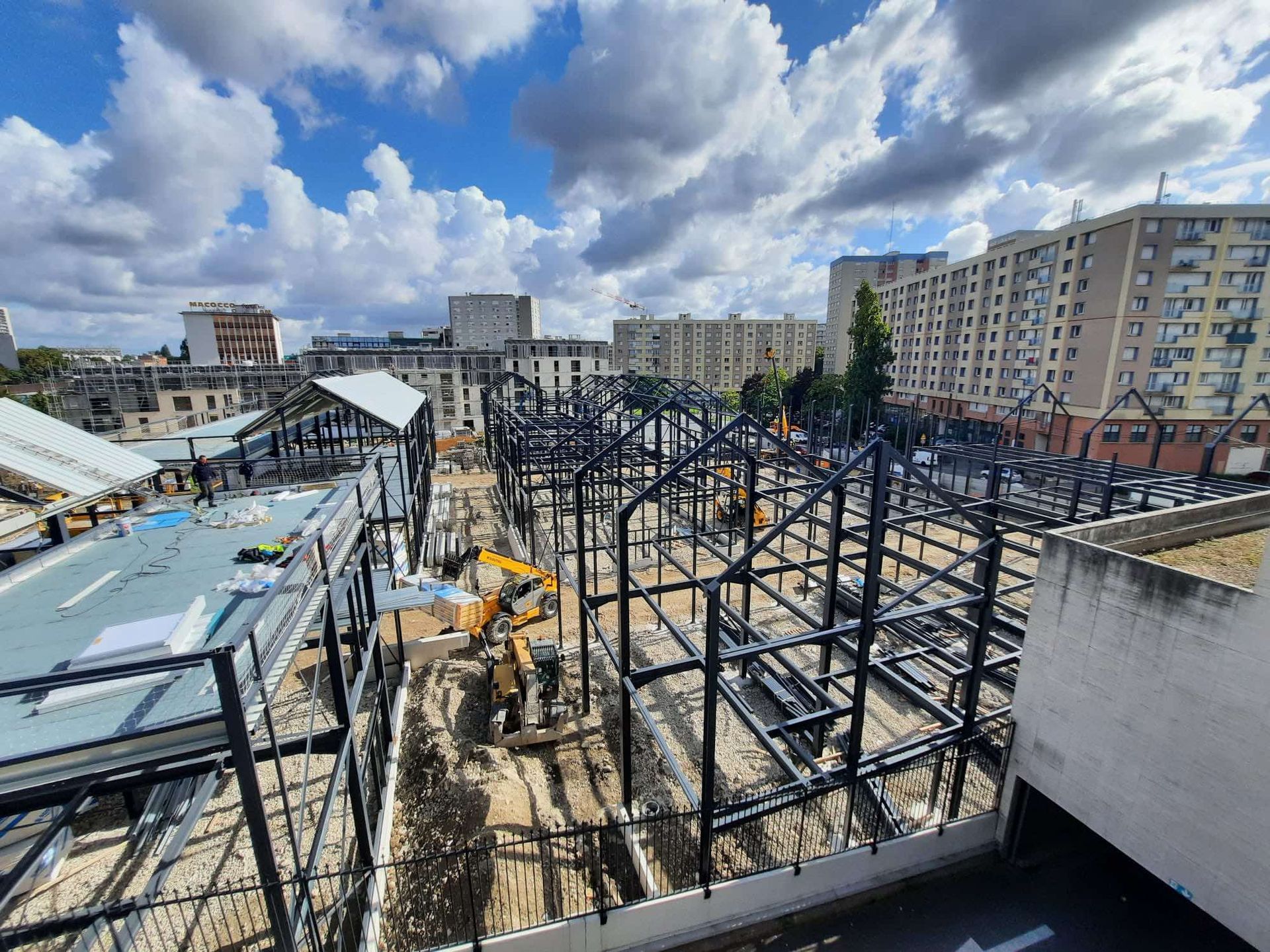 Chantier de construction d’une structure en acier sous un ciel nuageux, avec une excavatrice jaune et des immeubles résidentiels à proximité.