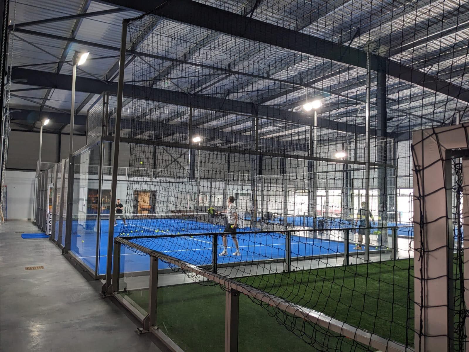 Complexe sportif couvert avec des terrains en gazon synthétique bleu, entourés de filets noirs et de clôtures métalliques
