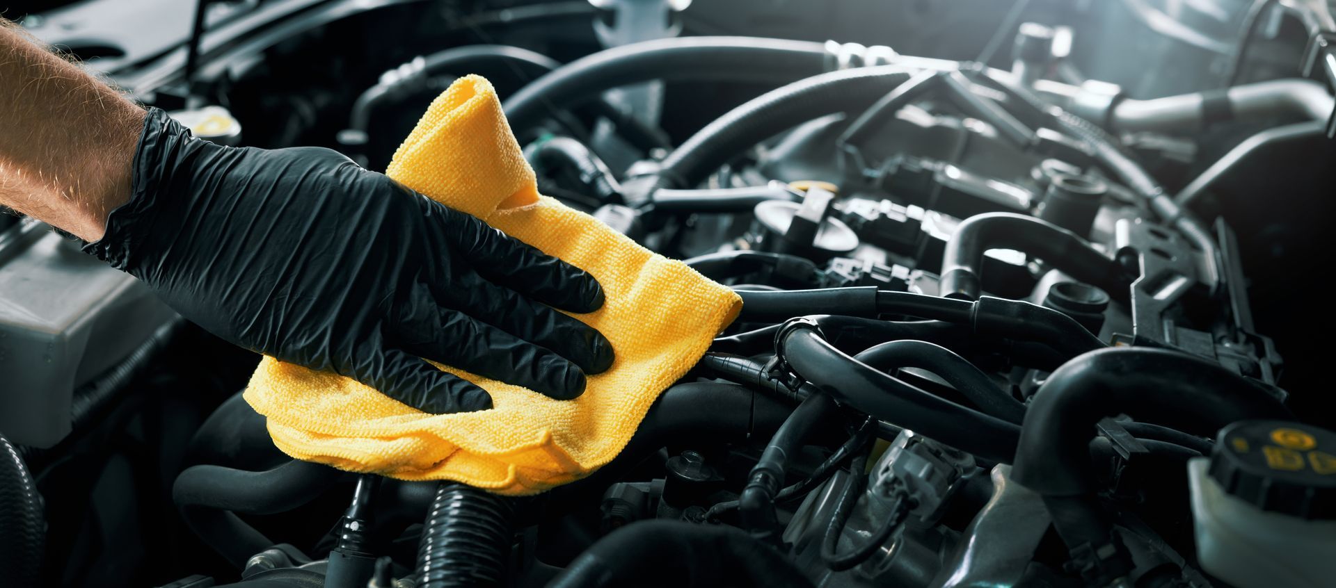 Une personne portant des gants noirs essuie le moteur d'une voiture avec un chiffon jaune.