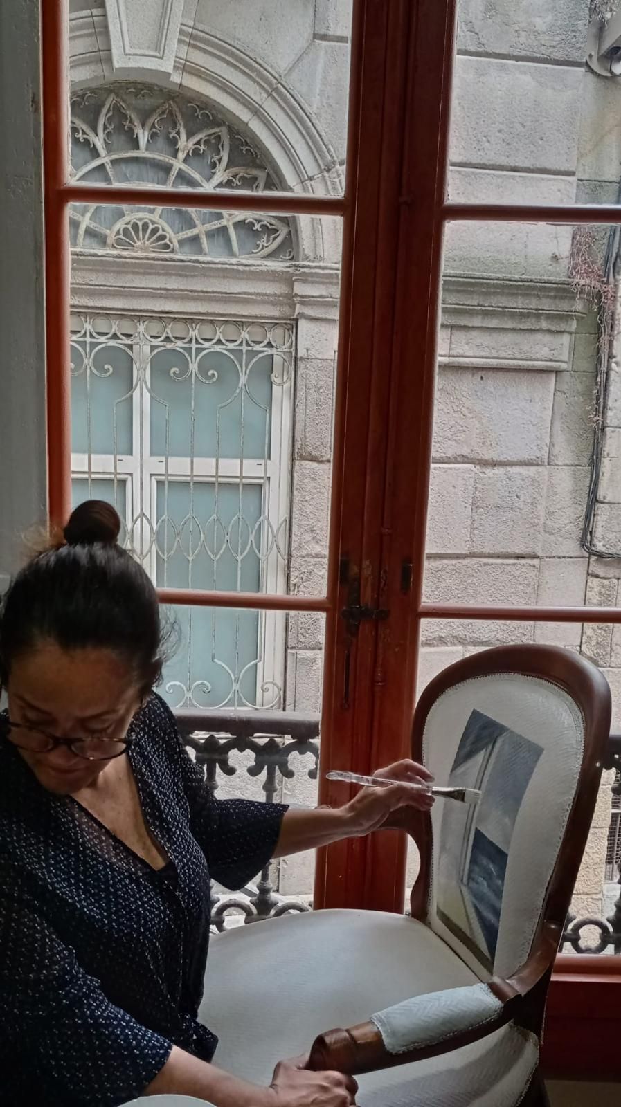 Mujer pintando una silla frente a una ventana, con gafas y una impresión en blanco y negro en la parte superior. Edificio ornamentado en el exterior.