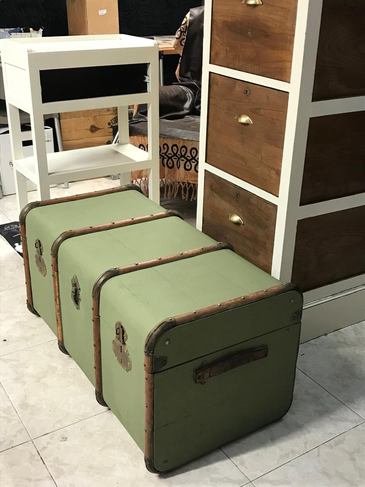 Baúles de almacenamiento verdes con detalles marrones frente a una cómoda blanca y marrón, mesita de noche en una tienda.