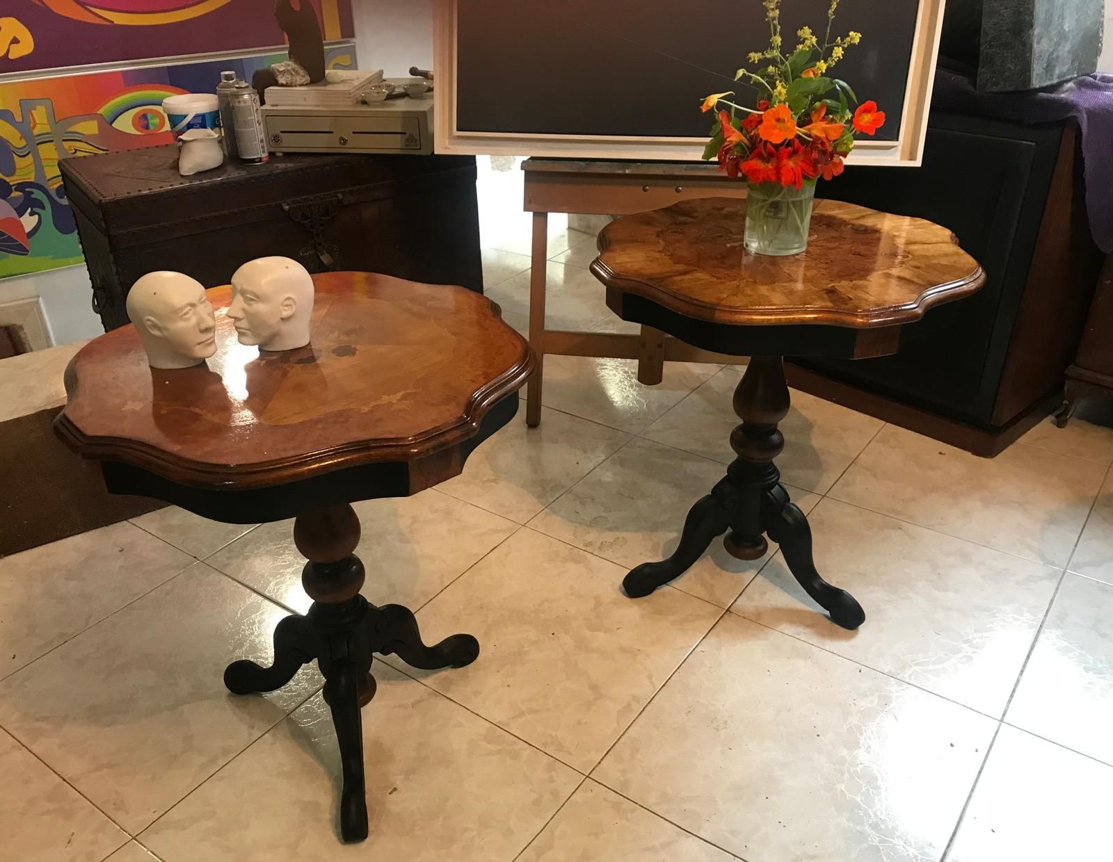 Dos mesas auxiliares ornamentadas con patas oscuras. Una tiene una escultura y la otra flores, en una habitación con obras de arte y muebles vintage.