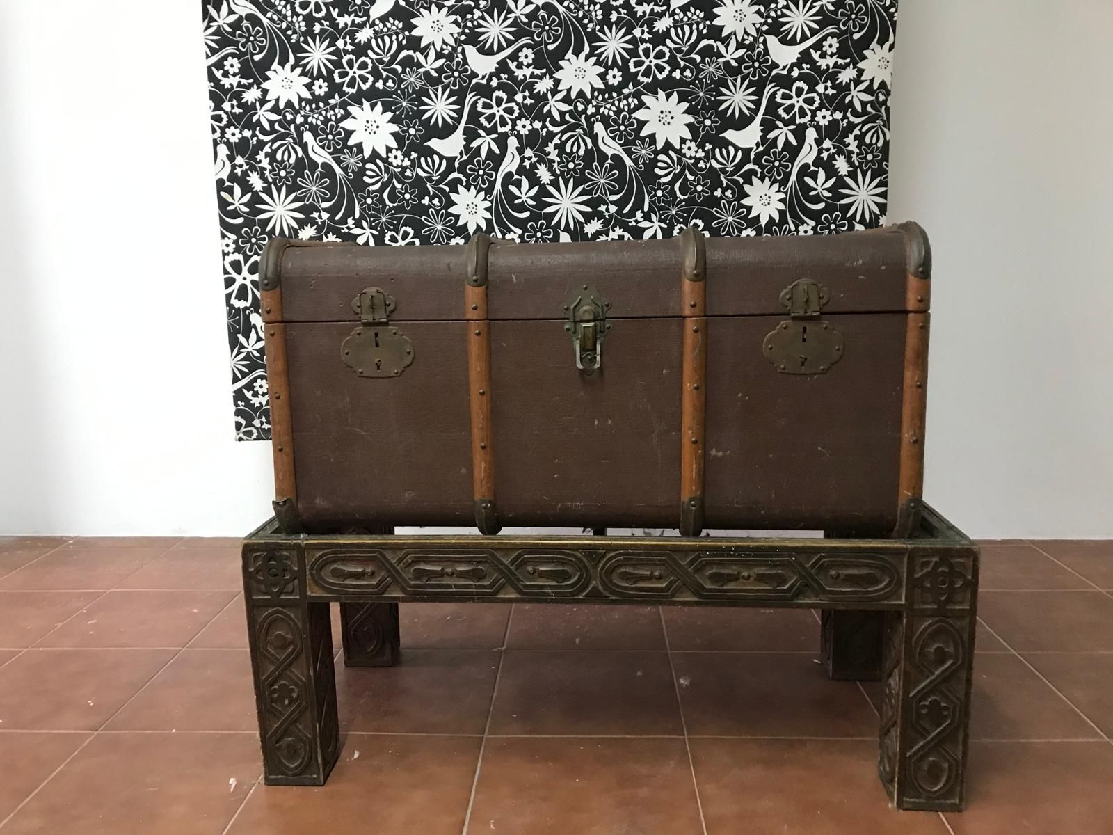 Baúl marrón antiguo sobre una mesa decorativa, sobre un fondo con estampado floral en blanco y negro.