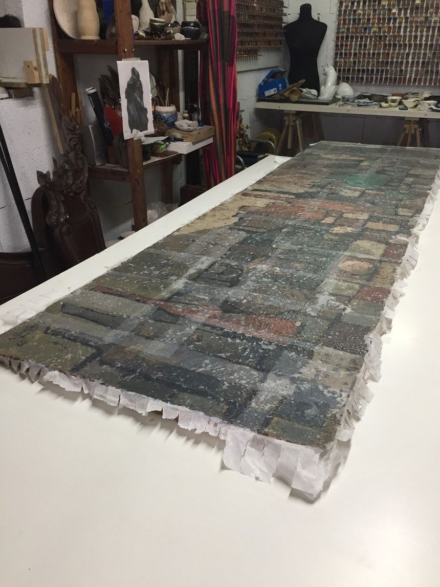 Obra de arte rectangular de gran tamaño sobre una mesa blanca, con una textura similar al ladrillo. La obra tiene un borde claro.