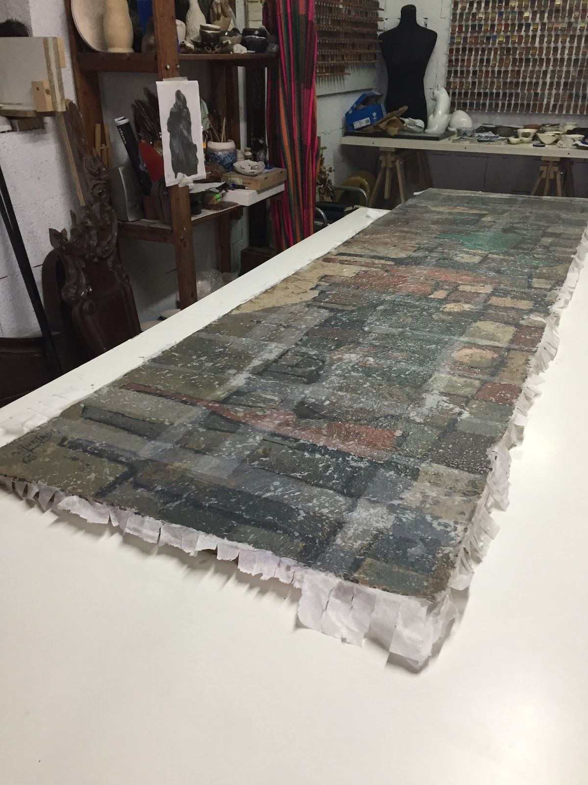 Obra de arte alargada y rectangular, con una superficie colorida y texturizada, que reposa sobre una mesa blanca en un estudio.