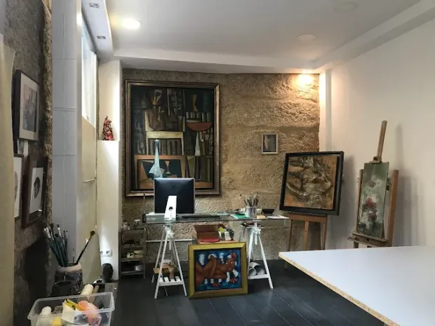 Un estudio de arte con cuadros, caballetes y un escritorio. Un gran cuadro cuelga de una pared de piedra, con materiales de pintura y una computadora cerca.