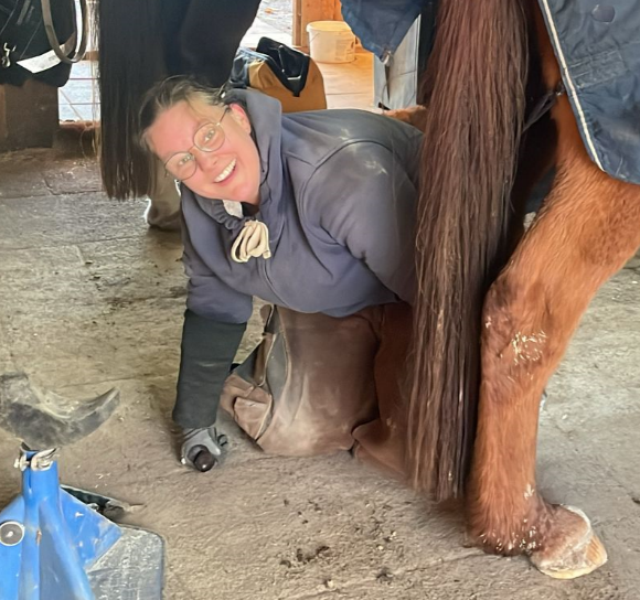 Eine Person mit Brille und grauem Kapuzenpullover kniet in einem Stall und schneidet mit einem Handwerkzeug den Huf eines Pferdes.