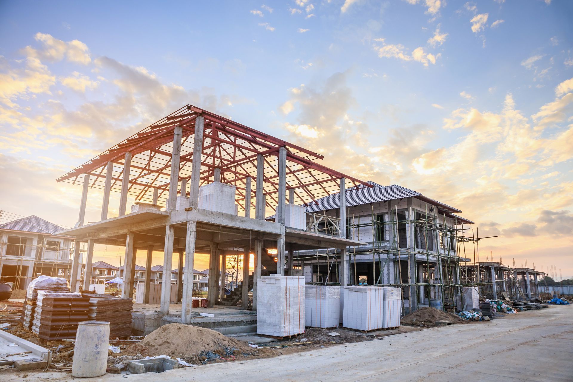 Chantier de construction avec maisons inachevées, charpentes en béton et ciel de coucher de soleil.