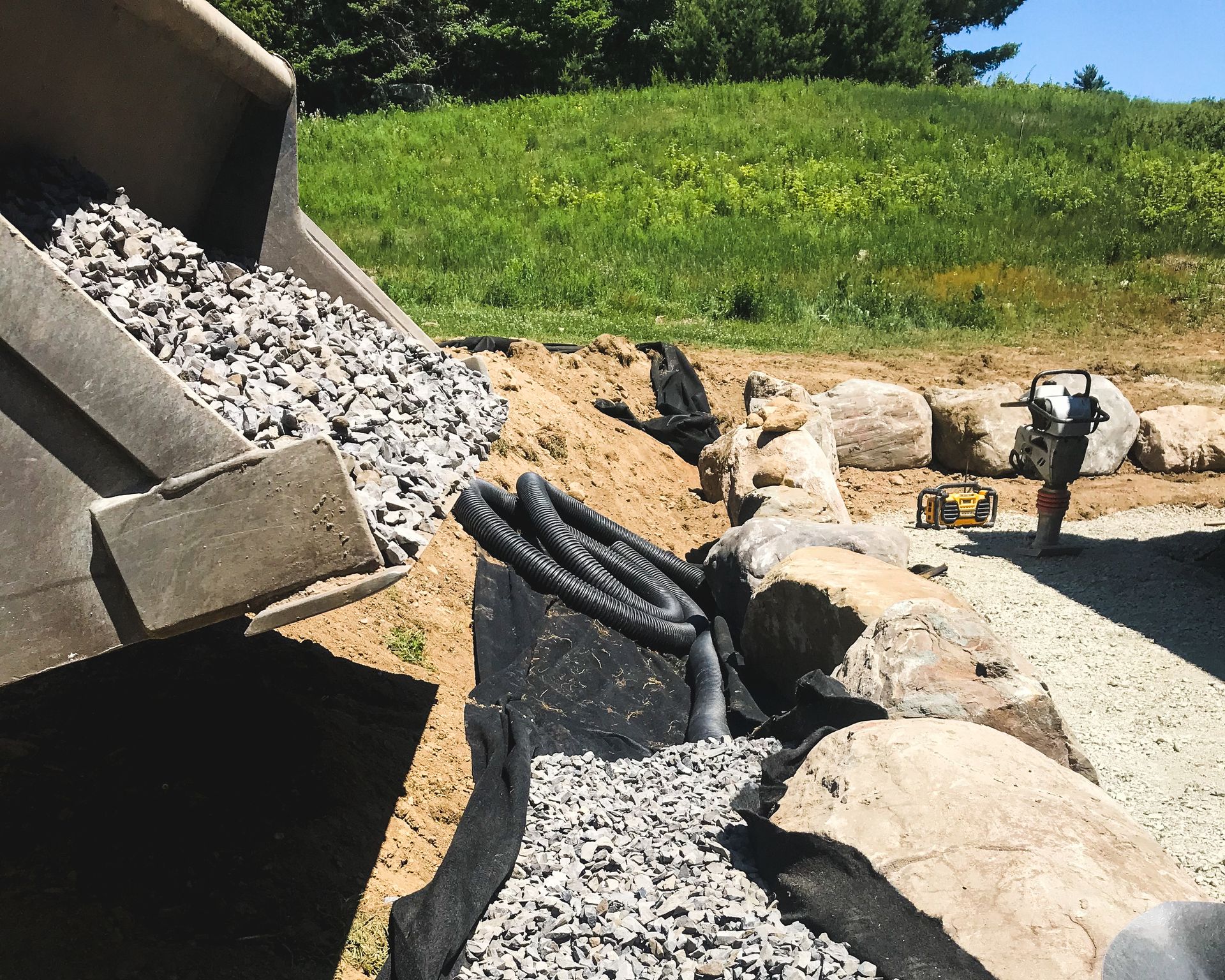 Un chantier de construction montrant une pelleteuse déversant du gravier. De gros rochers et des tuyaux de drainage se trouvent à proximité.
