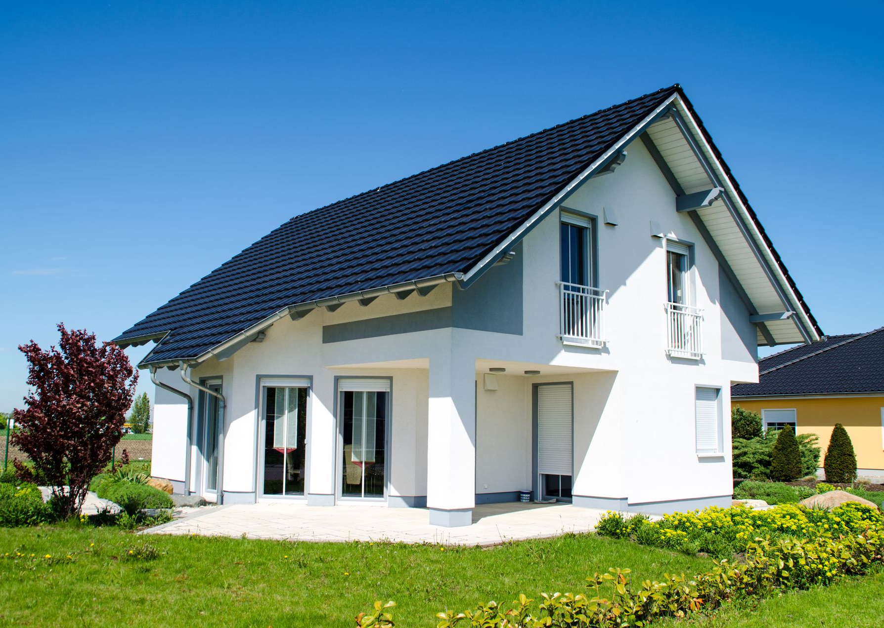 Maison blanche à deux étages avec toit sombre, fenêtres et cour verte sous un ciel bleu.