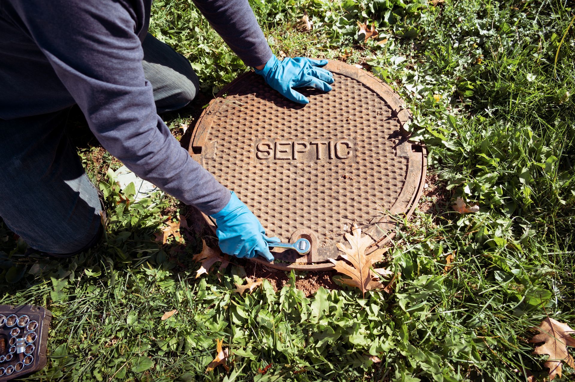 Une personne portant des gants bleus ouvre le couvercle d'une fosse septique à l'extérieur dans l'herbe.
