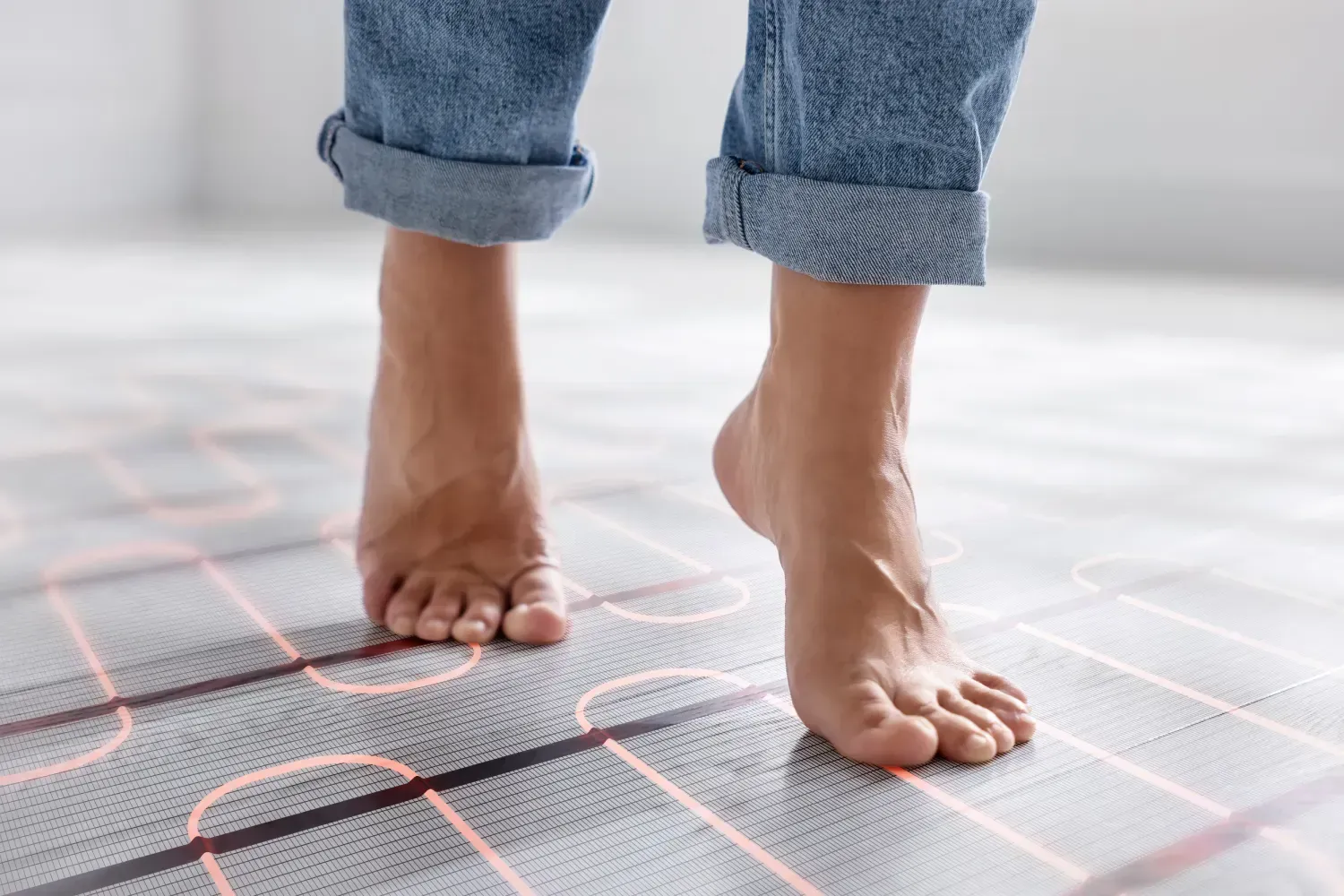 Pies descalzos caminando sobre una estera gris de calefacción por suelo radiante con bobinas eléctricas naranjas visibles.