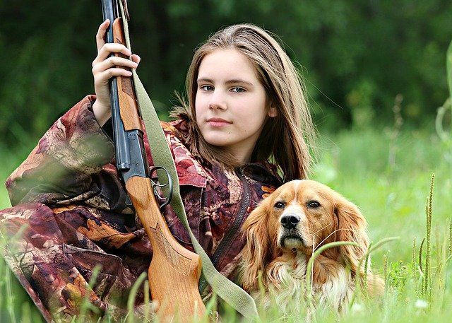 Chica con camuflaje sosteniendo un rifle, sentada en el césped con un spaniel.