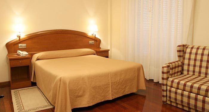 Dormitorio de hotel con una cama, mesitas de noche, un sillón estampado y una ventana con persianas.