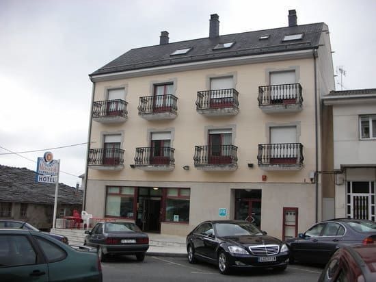 Un edificio de tres pisos con balcones, tienda debajo y autos estacionados al frente.