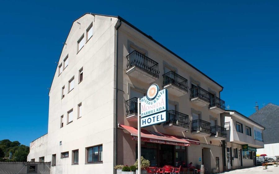 Exterior del hotel con balcones, un cartel y un toldo rojo. Luz solar brillante y cielo azul.