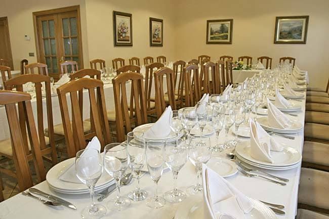 Comedor formal con mesas largas preparadas para comer. Mantelería, cubiertos y sillas blancas.