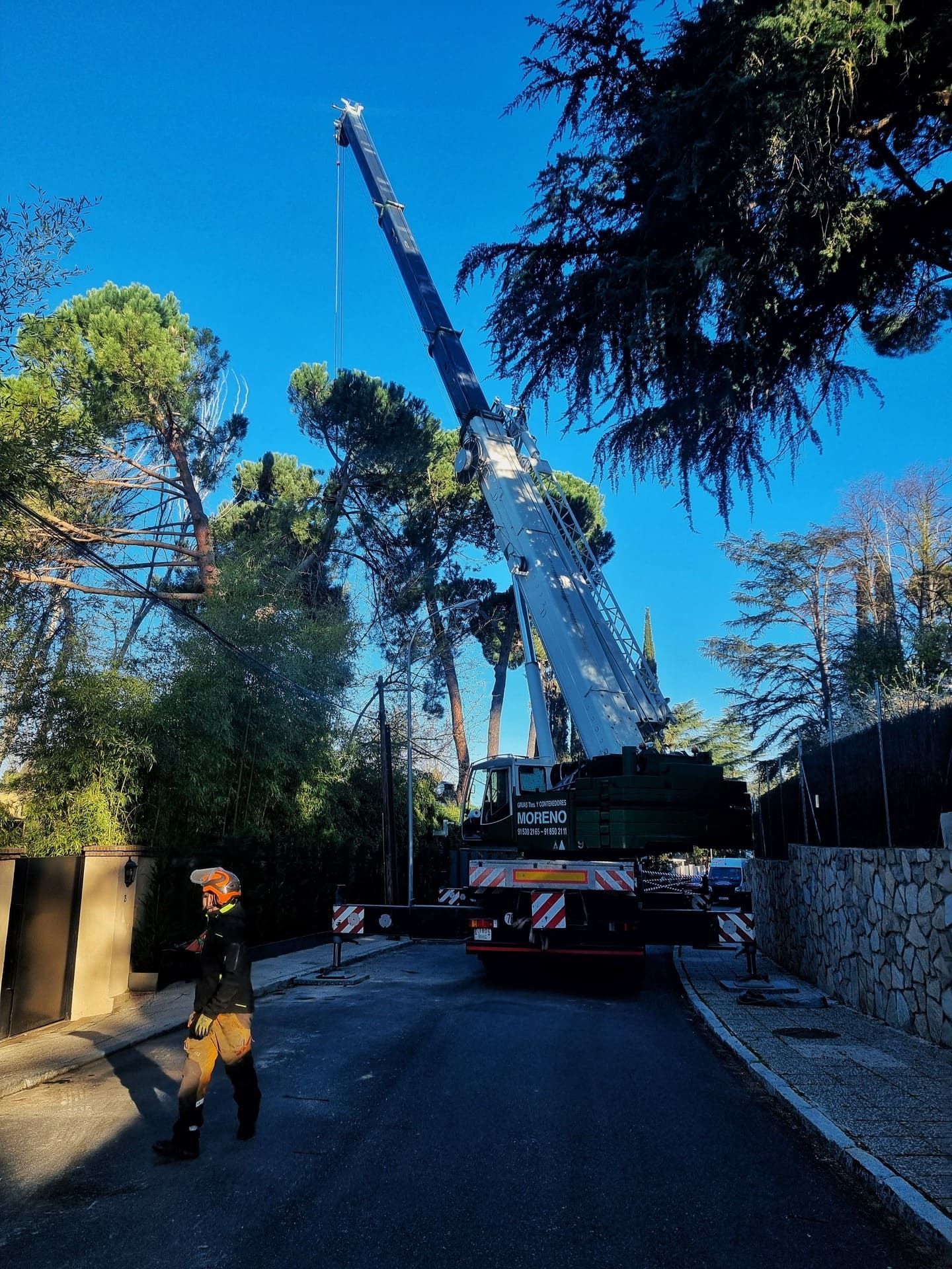 Una grúa levanta una gran viga en una carretera estrecha junto a árboles y un muro de piedra, con un trabajador con casco cerca.