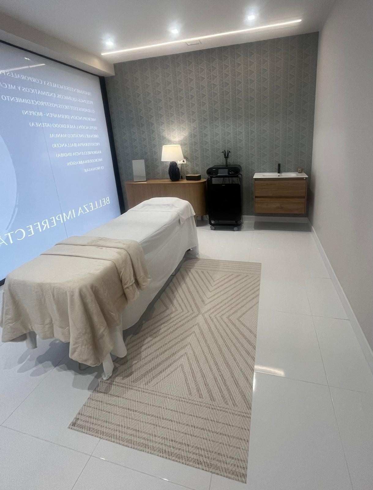 Sala de tratamiento minimalista con camilla de masaje cubierta, lámpara, alfombra y armario en tonos grises suaves.