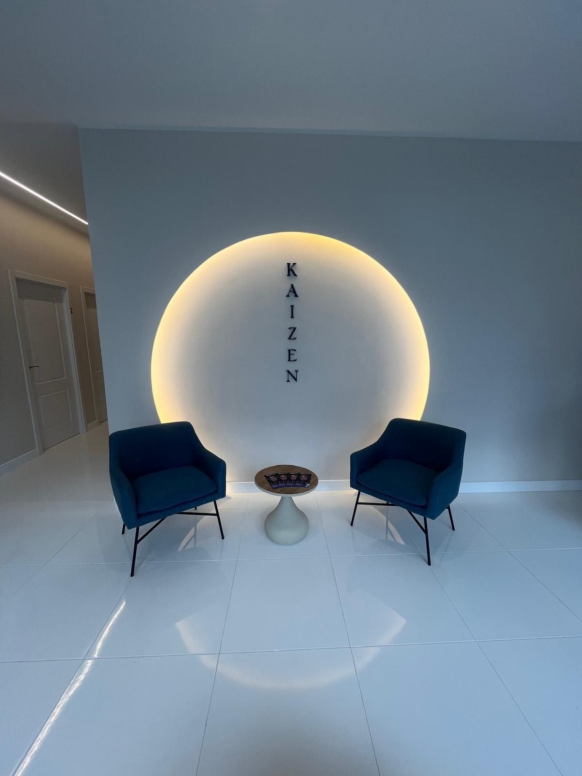 Salón moderno con dos sillas azules, una mesa pequeña y una lámpara de pared circular con luz tenue.