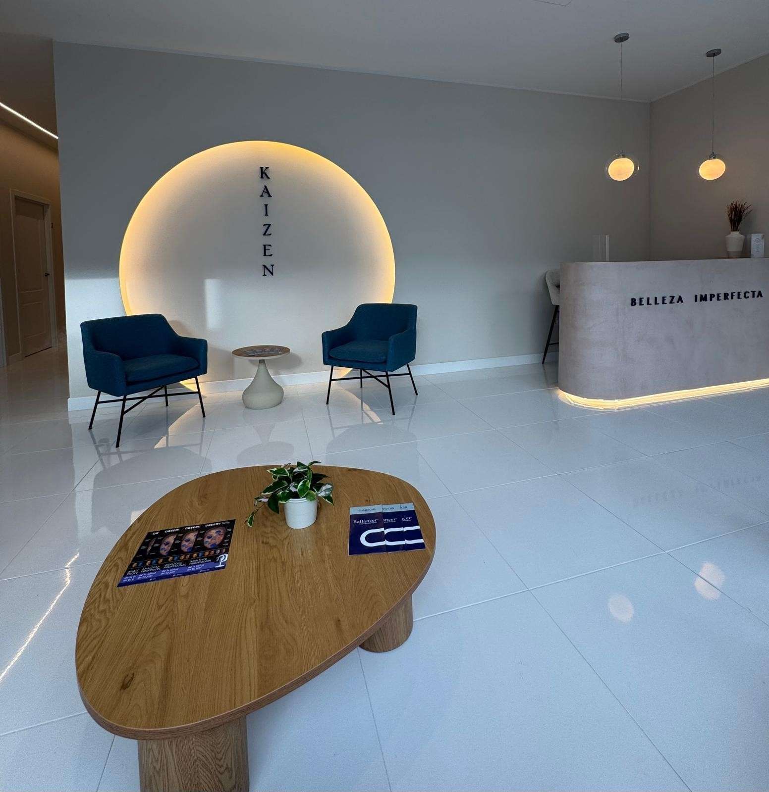 Vestíbulo moderno con sillas azules, mostrador de recepción blanco y mesa de centro de madera sobre un suelo brillante.