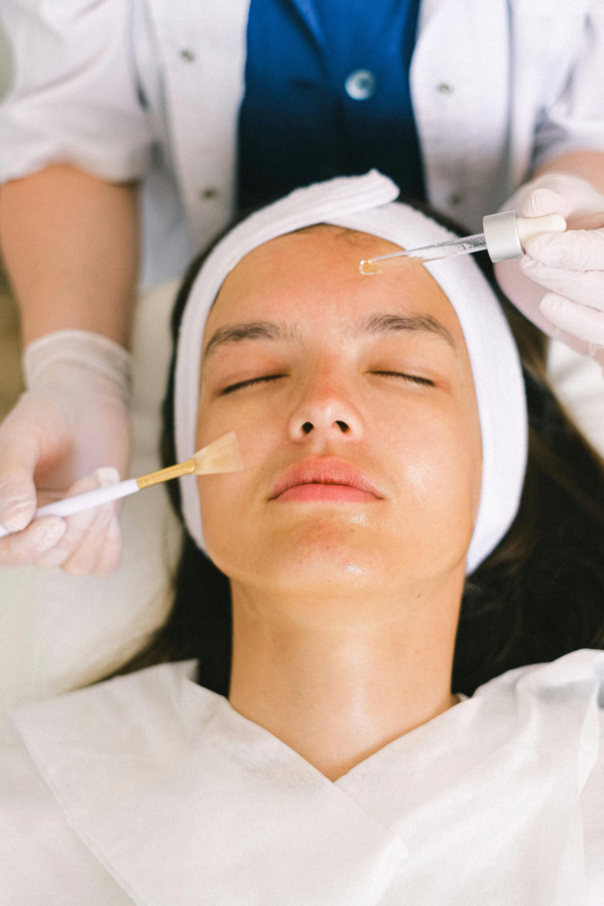 Una clienta recibe un tratamiento facial mientras está recostada con los ojos cerrados y un pañuelo en la cabeza.