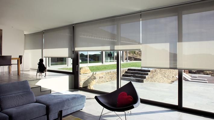 Interior de una casa moderna con grandes ventanales, cortinas enrollables de color gris transparente y muebles contemporáneos.