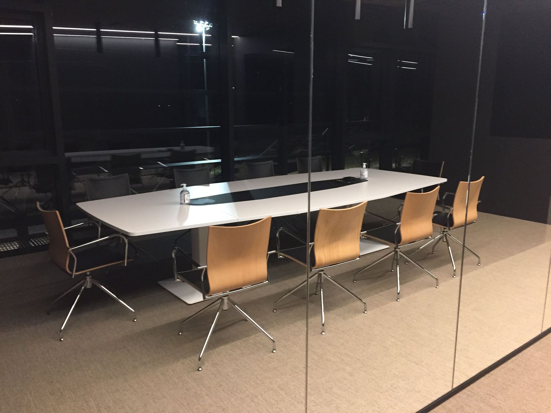 Sala de conferencias con mesa blanca y sillas beige, frente a una pared de cristal oscuro.