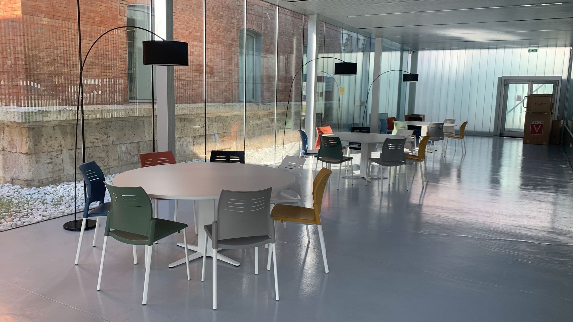 Habitación moderna con grandes ventanales, mesas redondas y sillas coloridas. El suelo brillante refleja la luz.