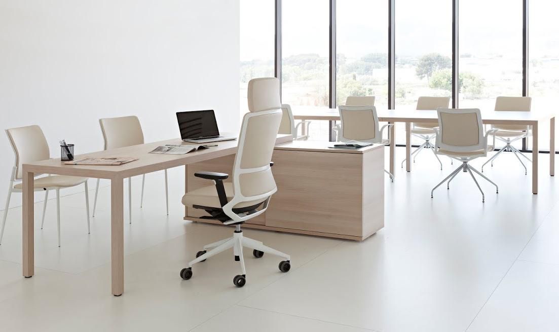 Oficina moderna con mesas largas, sillas y amplios ventanales. Hay una silla de escritorio frente a una computadora y varias sillas frente a la mesa.