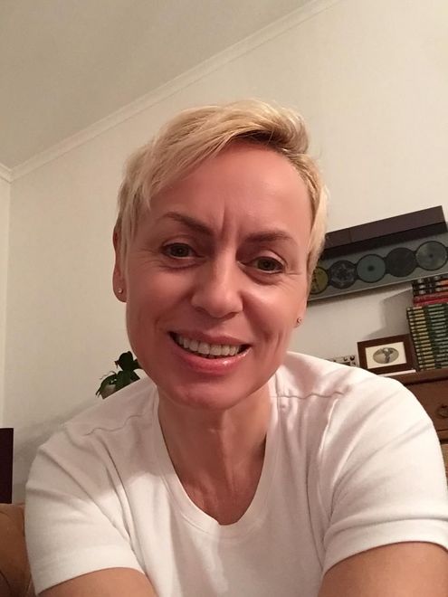 Persona sorridente in un ambiente interno con camicia bianca, capelli biondi corti e una parete beige alle sue spalle.