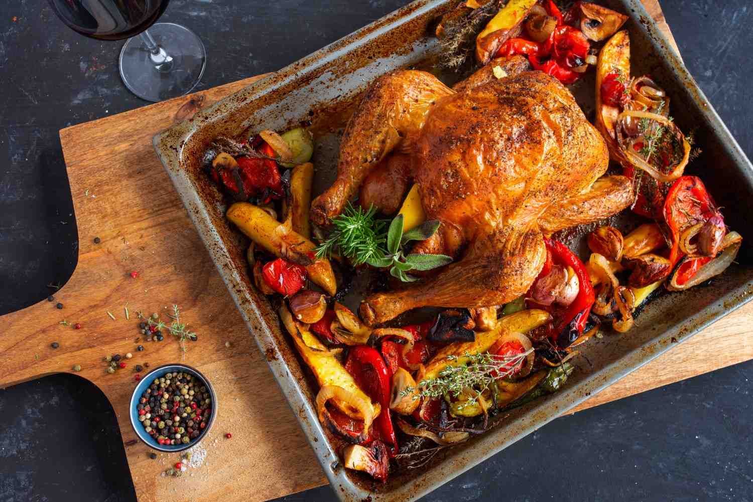 Pollo asado en una bandeja con verduras y hierbas, sobre una tabla de madera con un cuenco de granos de pimienta.