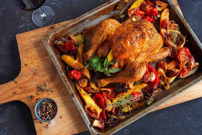 Pollo asado en una bandeja con verduras y hierbas, sobre una tabla de madera con un cuenco de granos de pimienta.