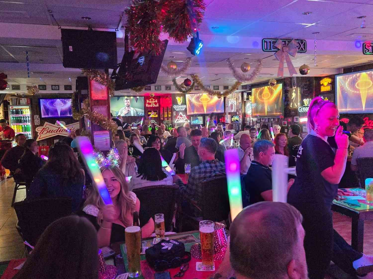 Un bar abarrotado de gente sentada en mesas, luces de neón y una cantante actuando a la derecha.