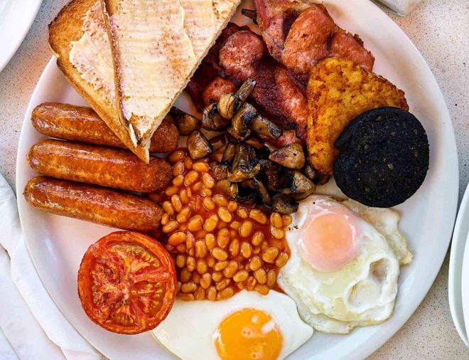 Desayuno inglés completo con salchichas, tocino, huevos, frijoles, tomate, champiñones, tostadas y morcilla en un plato blanco.