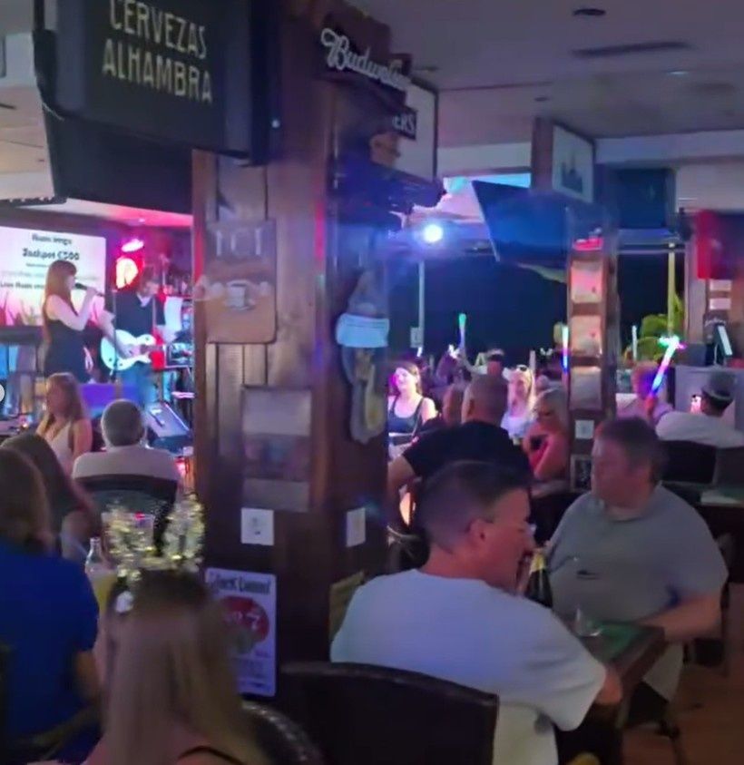 Patio de bar abarrotado con gente sentada cerca de un escenario de música en vivo bajo luces de colores.