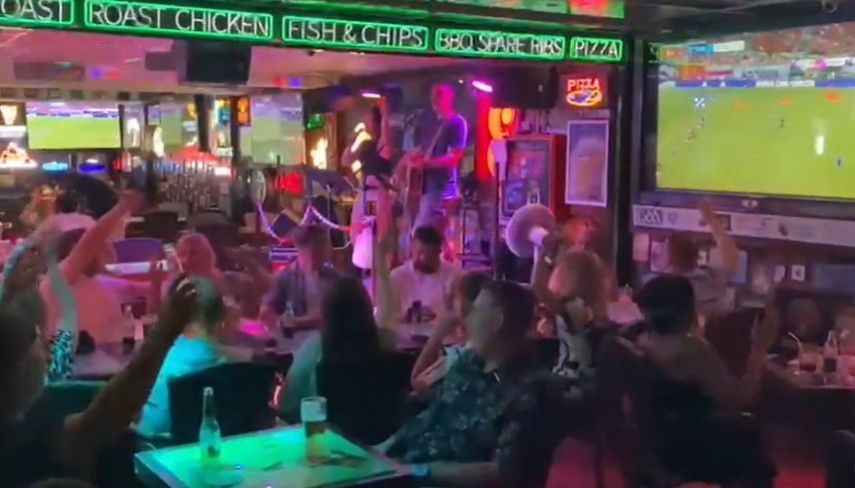 Bar deportivo abarrotado de gente, iluminado con luces de neón, viendo un partido de fútbol en una gran pantalla de televisión.