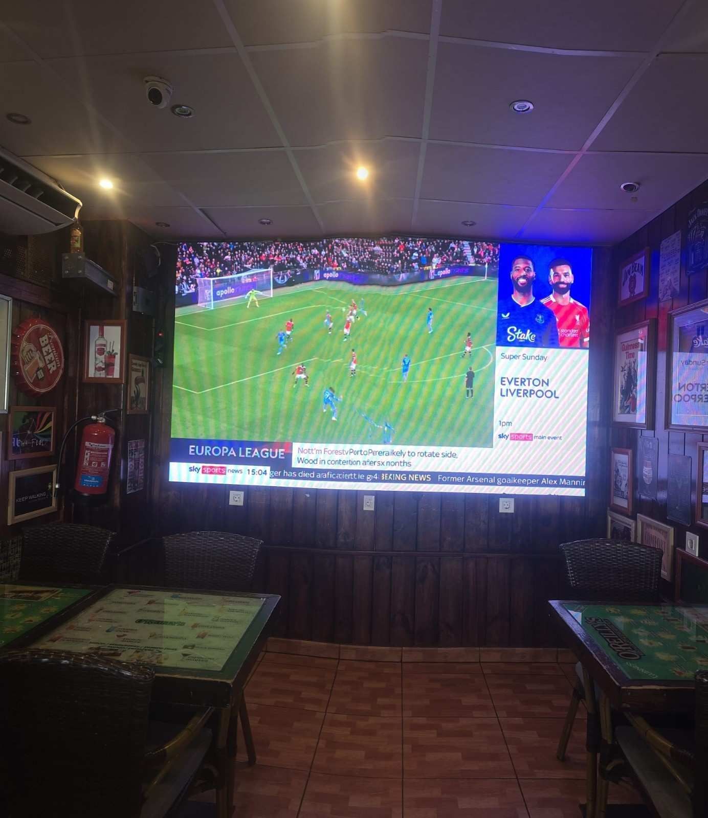 Bar deportivo con una gran televisión que retransmite un partido de fútbol, ​​flanqueado por mesas y con una decoración de pared tenue.