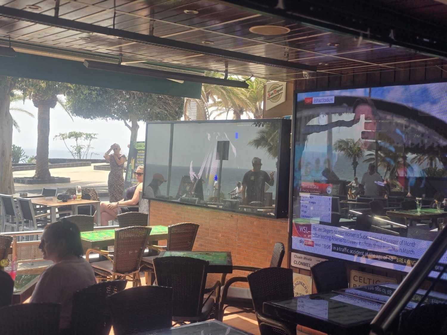 Cafetería al aire libre con mamparas de cristal, sillas de mimbre y vistas al mar bajo un techo que proporciona sombra.