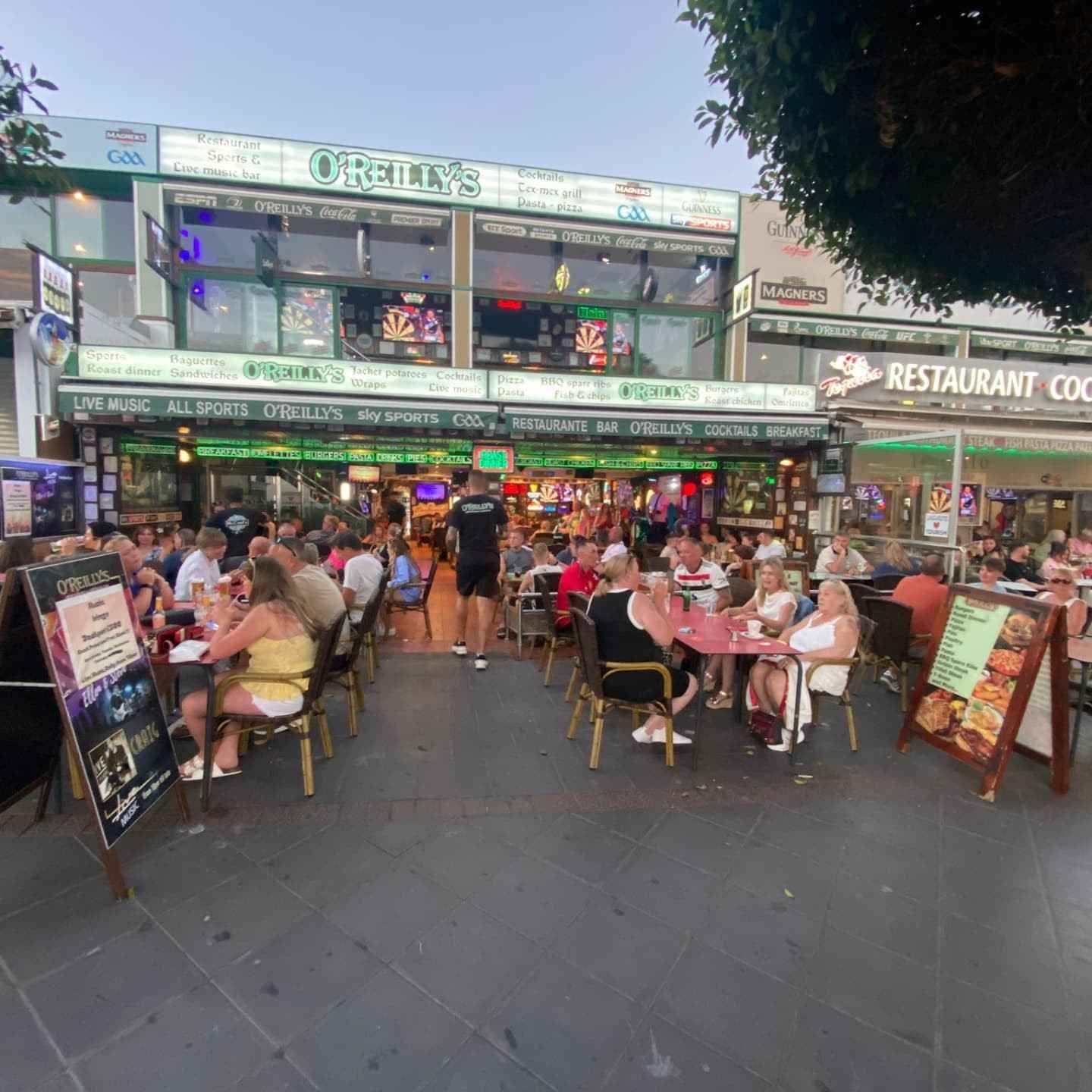 Terraza de restaurante al aire libre repleta de comensales bajo luces de colores al atardecer.