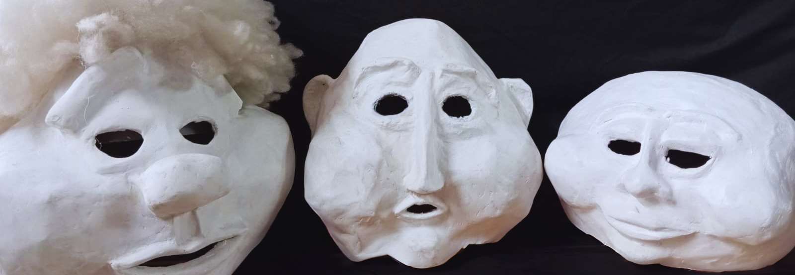 Tres máscaras de papel maché blanco con diferentes expresiones faciales, dispuestas sobre una superficie negra.