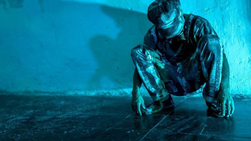 Una persona se agacha en un suelo oscuro, con ropa brillante y holgada. Una luz azul proyecta sombras en la pared.